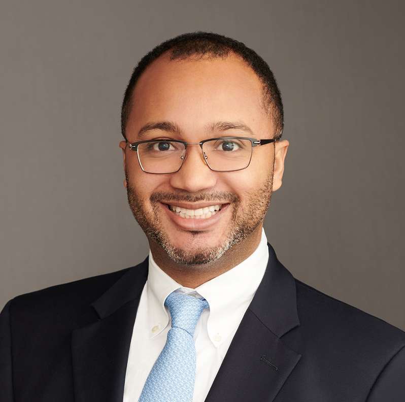 Aleksandr Spencer smiling, wearing glasses, a dark suit, white shirt, and blue tie against a plain gray background.