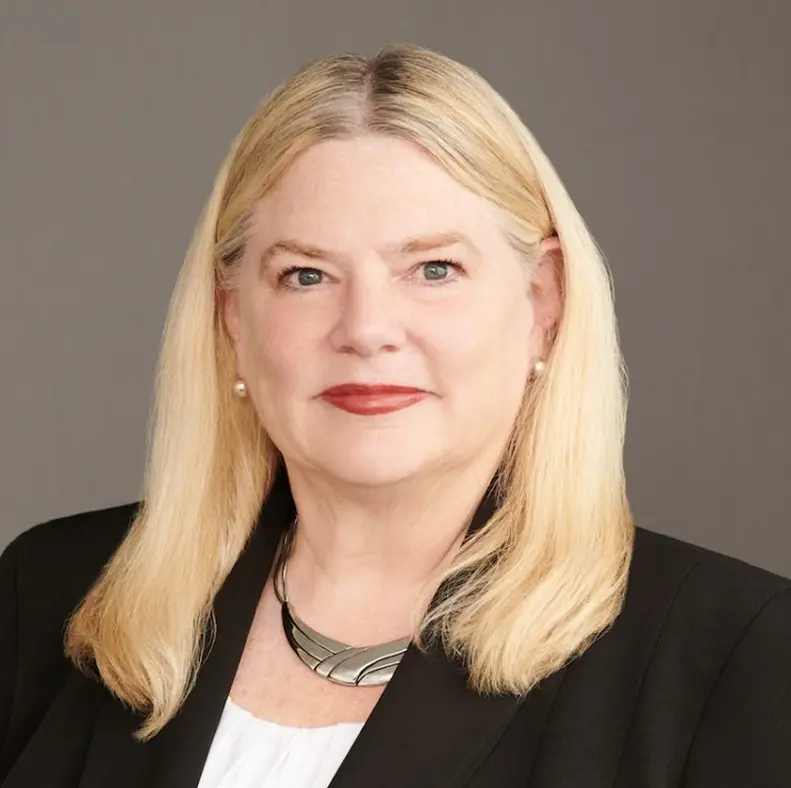 Woman with long blonde hair, wearing a black blazer and silver necklace, facing forward against a gray background.