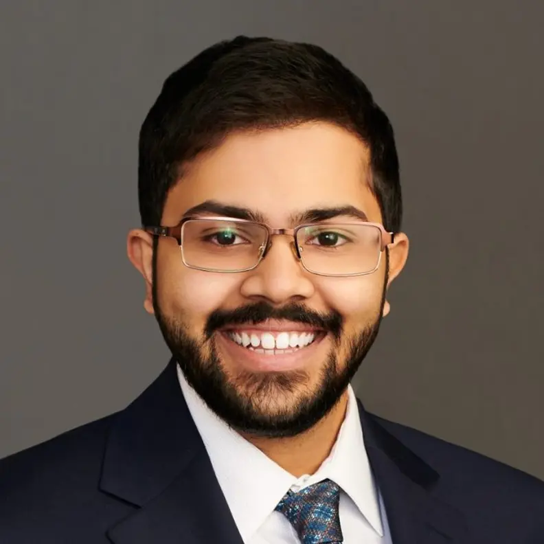 Smiling Shesh Batni in a suit, wearing glasses, with short hair and a beard, against a plain background.