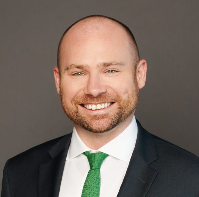 Smiling man with a beard in a suit and green tie, against a gray background.