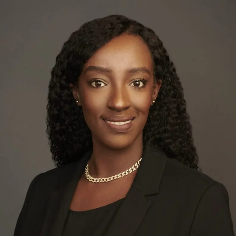 Saro Gahima smiles in a professional headshot, wearing a black blazer and a gold necklace, against a neutral background.