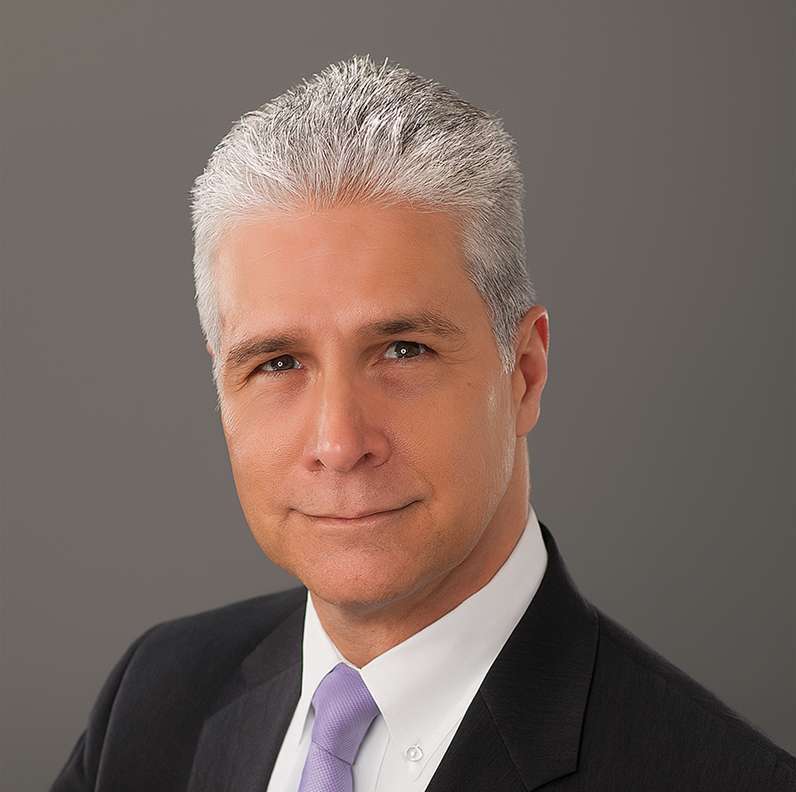 Man with silver hair in a suit and lavender tie, against a gray background.