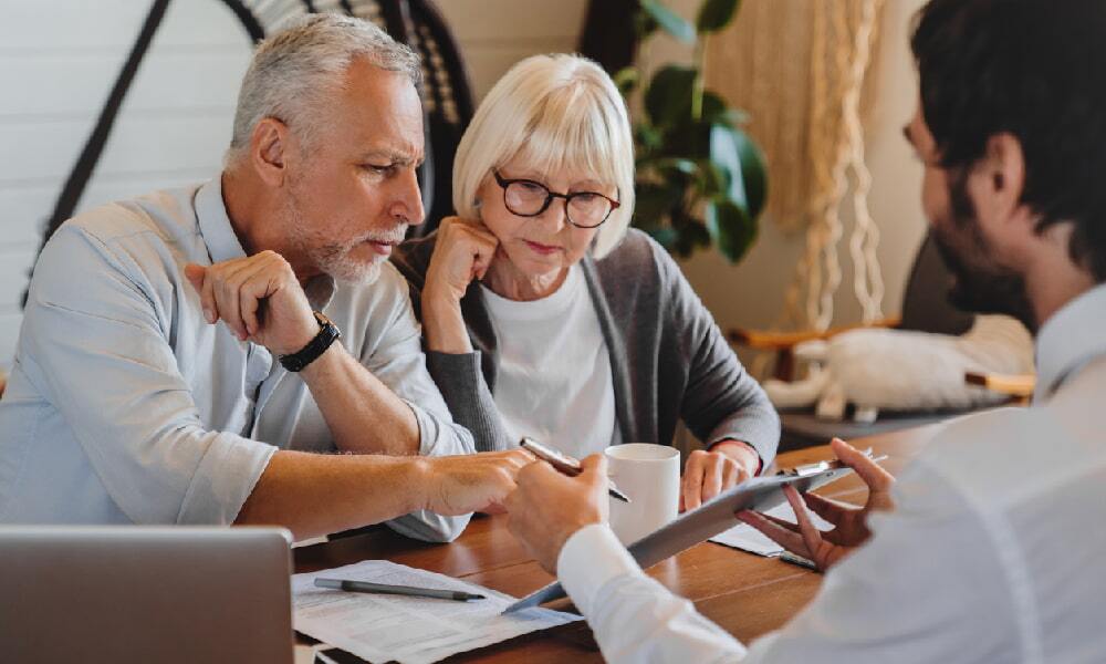 An elderly couple sits at a desk and looks at a tablet while talking with a financial advisor about avoiding retirement mistakes.